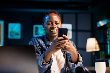 Positive african american blogger working remotely from home late at night while texting on her...