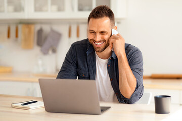Gig economy concept. Young positive man freelancer working remotely at home, looking at laptop and consulting client via cellphone, sitting at kitchen