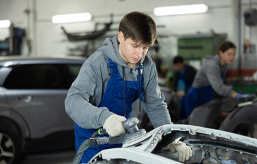 Male mechanic prepares car for painting in workshop - using grinding machine he sands the bumper before painting