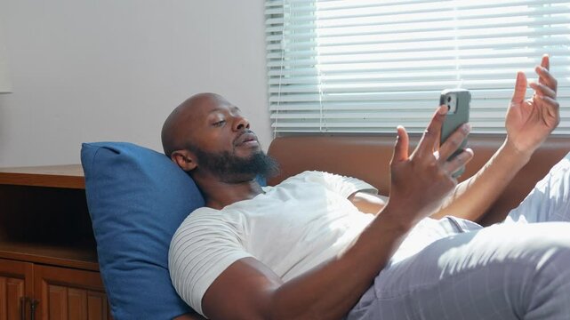 Young african american male lounging on living room sofa, scrolling smartphone with growing boredom, gradually succumbing to drowsiness and drifting into unintended nap