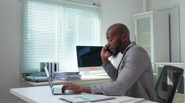Professional businessman multitasking while working on laptop and communicating via phone, efficiently managing corporate tasks in bright, modern office setting