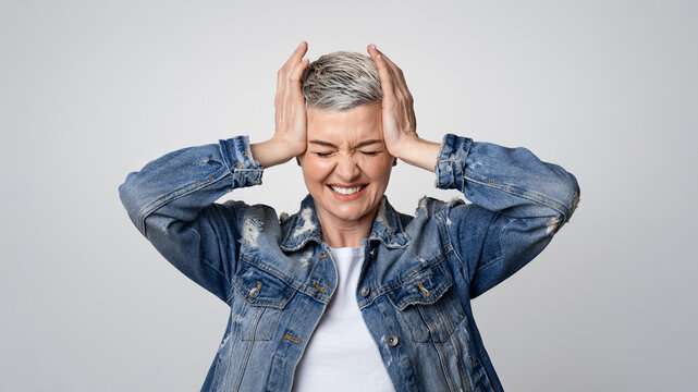 Headache Concept. Sick mature woman touching her temples, suffering from migraine or stress, emotionally reacting to acute pain, light background