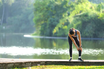 Young sporty asian woman massaging her calf while dealing with a leg injury after an intense run in a public park near the serene lake, embracing an active lifestyle amidst nature © crizzystudio