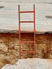 A red metal ladder positioned against the edge of a deep trench at a construction site. The exposed soil layers and broken asphalt indicate ongoing excavation work. © yulikolibrik