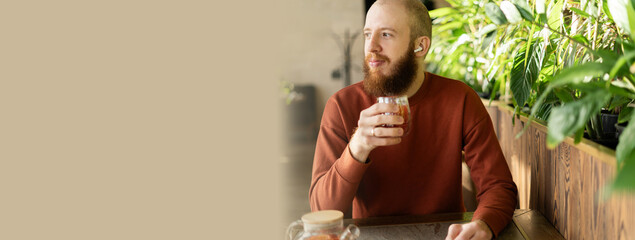 Bearded man in rust-colored sweater drinking herbal tea in cozy cafe surrounded by lush green...