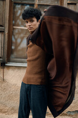 Obraz premium Fashionable young man in brown sweater and jeans standing in front of old building with arms outstretched, brown coat draped over shoulders