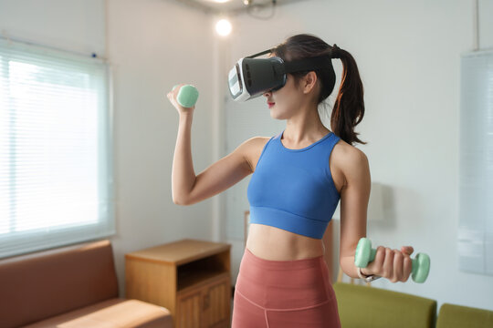 Young woman wearing sportswear is lifting weights while using a virtual reality headset in her living room, experiencing an immersive fitness workout