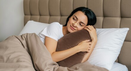 Naklejka premium A young woman with long dark hair rests on a comfortable bed, smiling while embracing a soft pillow. The scene is cozy, featuring neutral tones and soft bedding. Comfort and relaxation are evident.