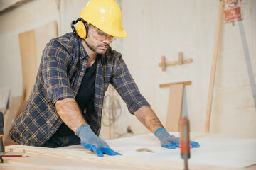 Young carpenter in a yellow helmet, blue gloves, and plaid shirt using a circular saw to cut wood in a furniture factory. Ideal for showcasing woodworking skills and construction techniques. Labor day