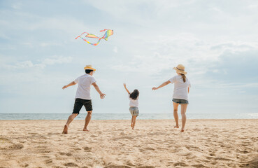 Asian family is flying a kite high in the sky while running on the beach. Happy parents mother and father with their child playing with kite, family is enjoying their time together, summer day