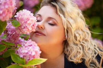 Fototapeta premium Portrait of a beautiful fat girl in a blue dress, against the background of a wall of live growing pink gartenias. The plus size woman enjoys the beauty of flowers