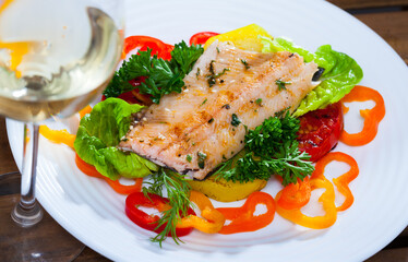 Roasted fillet of trout on cushion of fresh and baked vegetables and greens served on white plate with glass of wine