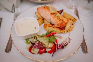 A plated meal featuring grilled salmon with lemon, roasted potatoes, fresh salad, and a side of creamy sauce, served on an elegant white dish..