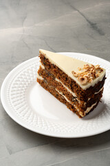 Carrot cake slice on white plate with frosting and nuts against a textured gray background