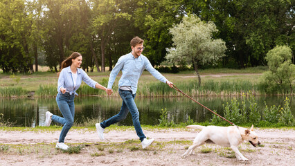 Active Pet Concept. Laughing couple chasing their running dog in park near lake, copy space, full...