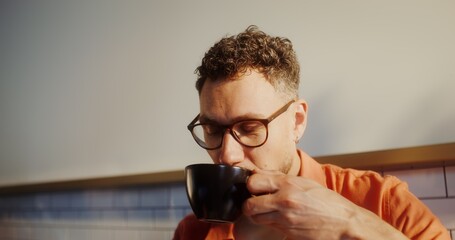 A stylish young man with glasses drinks coffee sitting in a cafe, face close-up
