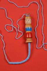 A top-down shot of a wooden doll with metal knitting hooks, adorned with blue yarn and a partially knitted cord against a red background © nahhan