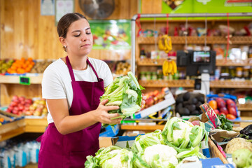 Supermarket female employee lays out fresh cauliflower on shelves in greengrocery store