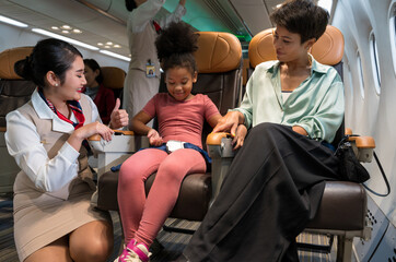 Female flight attendant in uniform kneeling beside young girl in pink clothing seated on aircraft...