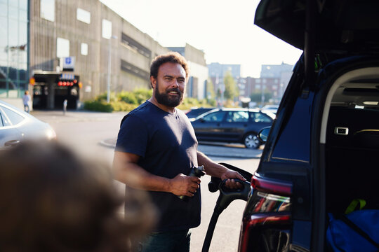 Bearded man charging electric car with plug at station on street