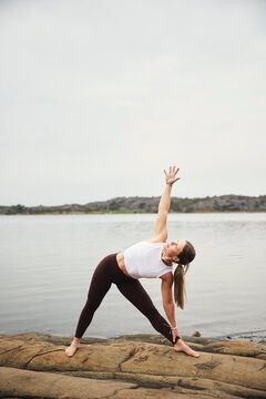 Full length of woman practicing Parivritta Trikonasana pose on rocks by river