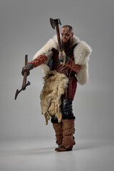 Warrior gripping two axes with intense gaze, man dressed in armor and fur cloak enhancing battle-ready stance against grey studio background. Concept of historical portrait. Rugged Viking aesthetic