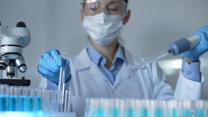A female scientist, wearing a mask, blue gloves and safety glasses, is using a micropipette to drop...