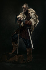 Obraz premium Viking warrior in rugged fur cloak, with axe and sword, standing in dramatic lighting against dark background. Concept of warriors bravery, medieval strength, Norse legend, historical portrait