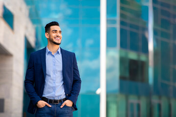 Fototapeta premium Handsome middle-eastern young businessman in stylish suit posing by office center, smiling arab entrepreneur having break after successful busienss meeting, standing on street, looking at copy space