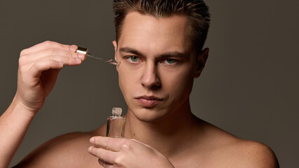 Obraz premium Shirtless man with serious expression applying serum with dropper near eye, looking directly at camera against brown studio background. Concept of selfcare and love, natural beauty, cosmetology.