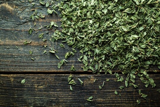 Overhead shot of dried thyme leaves scattered on rustic dark wooden surface, creating a natural and textured culinary background with muted green and brown tones.