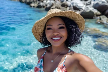 A beautiful Black woman taking a selfie at the beach, wearing a floral dress and straw hat and smiling. The clear blue water with rocks in the background on a sunny day