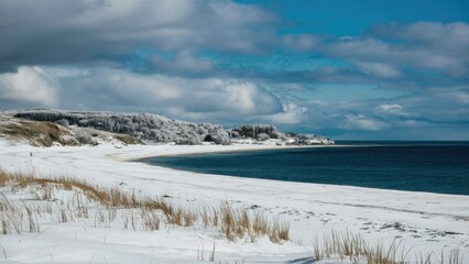 Snowy beach landscape with white sand covered in fresh snow contrasting against a turquoise sea under a partly cloudy sky with distant hills.