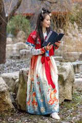 A young Asian woman in a traditional red Chinese costume walks in the park, reads national haiku...