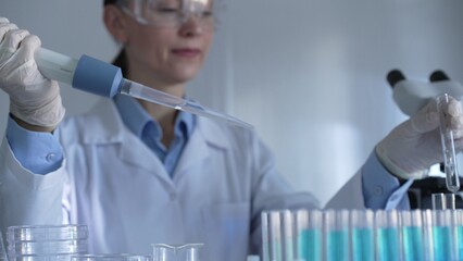 A scientist woman, wearing white gloves and safety glasses, is using a micropipette to drop a...