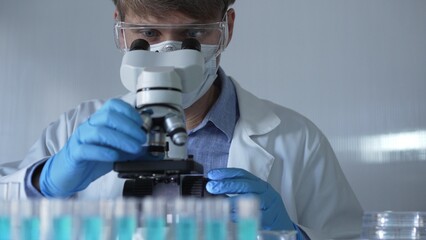 Scientist man, wearing a lab coat, mask, blue gloves and safety glasses, is using a binocular microscope surrounded by test tubes in laboratory. Portrait view. Medicine and science concept