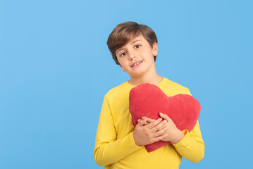 Charming young boy holding red heart in his arms in yellow sweater, isolated on blue background. Concept of love, compassion and good feelings