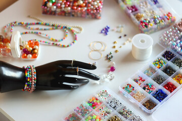 Hand figurine, colorful friendship bracelets, necklaces, various beads and DIY supplies on the table. Making jewelry at home. Selective focus.