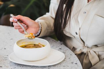 A close-up of a woman holding a spoonful of soup, seated at a stylish table. The bowl is white, and the soup contains various colorful ingredients.