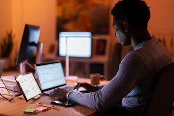 A young man wearing a light sweater sits at a desk in a dimly lit office, working on multiple devices including a laptop and tablet, with screens glowing in the background.