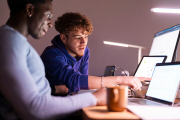 Two young professionals sit at a shared desk in a dimly lit office, reviewing stock market data displayed on multiple screens while discussing investment strategies.