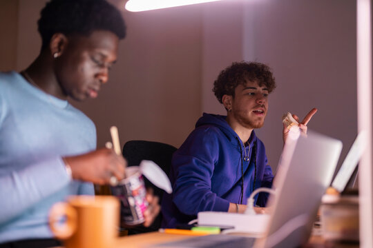 Two young men sit at a desk in a dimly lit workspace, one eating noodles with chopsticks and the other holding a sandwich while engaged in conversation.