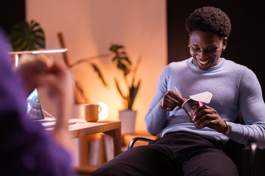 A young man in a blue sweater is laughing while eating instant noodles with chopsticks in a warmly lit workspace with a laptop and coffee mug nearby.