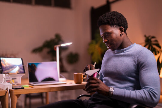 A young man in a blue sweater is laughing while eating instant noodles with chopsticks in a warmly lit workspace with a laptop and coffee mug nearby.