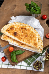 A golden-brown calzone rests on a wooden cutting board, surrounded by fresh basil leaves, cherry tomatoes, and a glass jar of mixed spices. A bottle of olive oil and a small knife are nearby