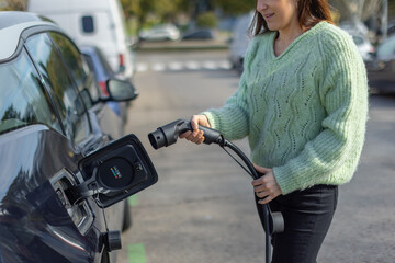 Cropped woman in a green sweater prepares to charge her electric vehicle, holding the charging cable near the port. Promoting eco-friendly transportation and clean energy solutions. Horizontal