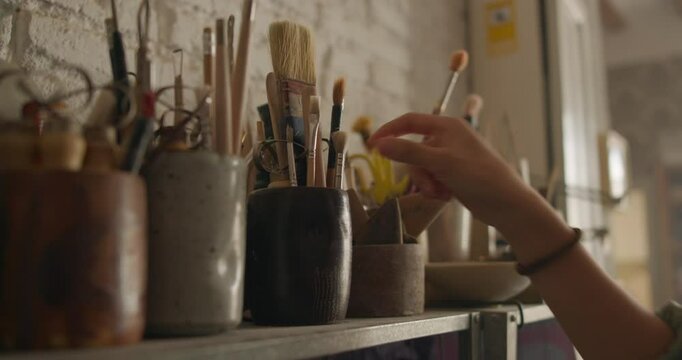 Artists hand selecting a paintbrush from a shelf filled with tools in a ceramic studio. Warm lighting and rustic setting enhance the creative atmosphere.