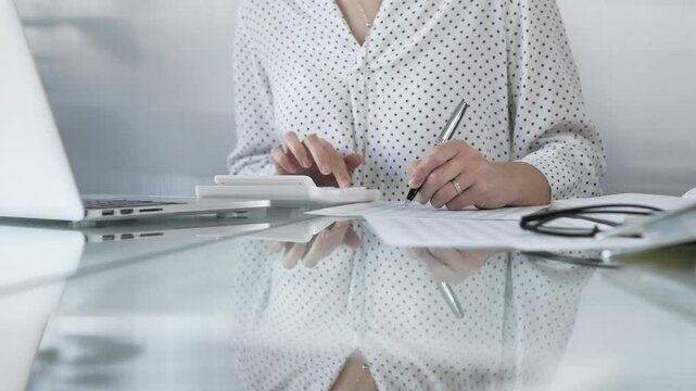 Focused businesswoman at a sleek glass desk in a contemporary office, using a calculator to crunch numbers and jotting down meticulous notes. Taxes, audit in business