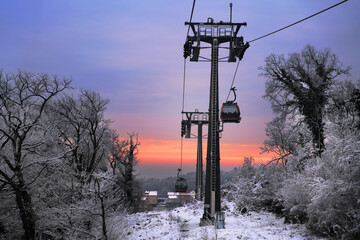 A cable car ascends into a snowy, tree-lined mountain landscape at twilight, with a vibrant sunset...