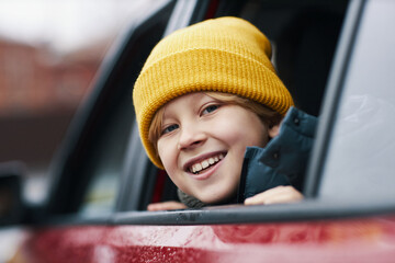 Smiling child with blonde hair wearing a bright yellow beanie, leaning out of car window, and...